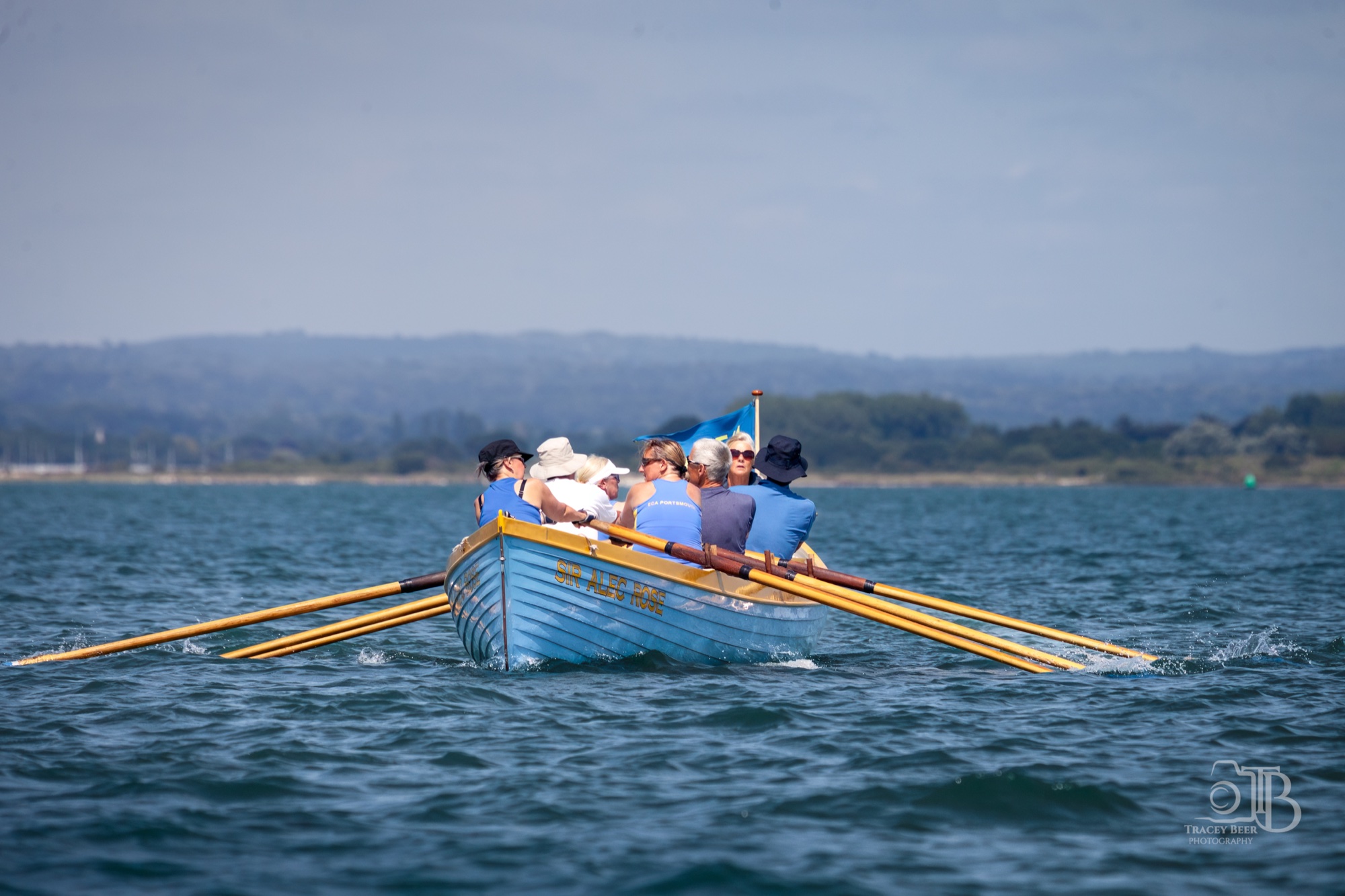 A crew rowing the blue fixed-seat boat Sir Alec Rose on calm harbour water, with a tree-lined shore beyond.
