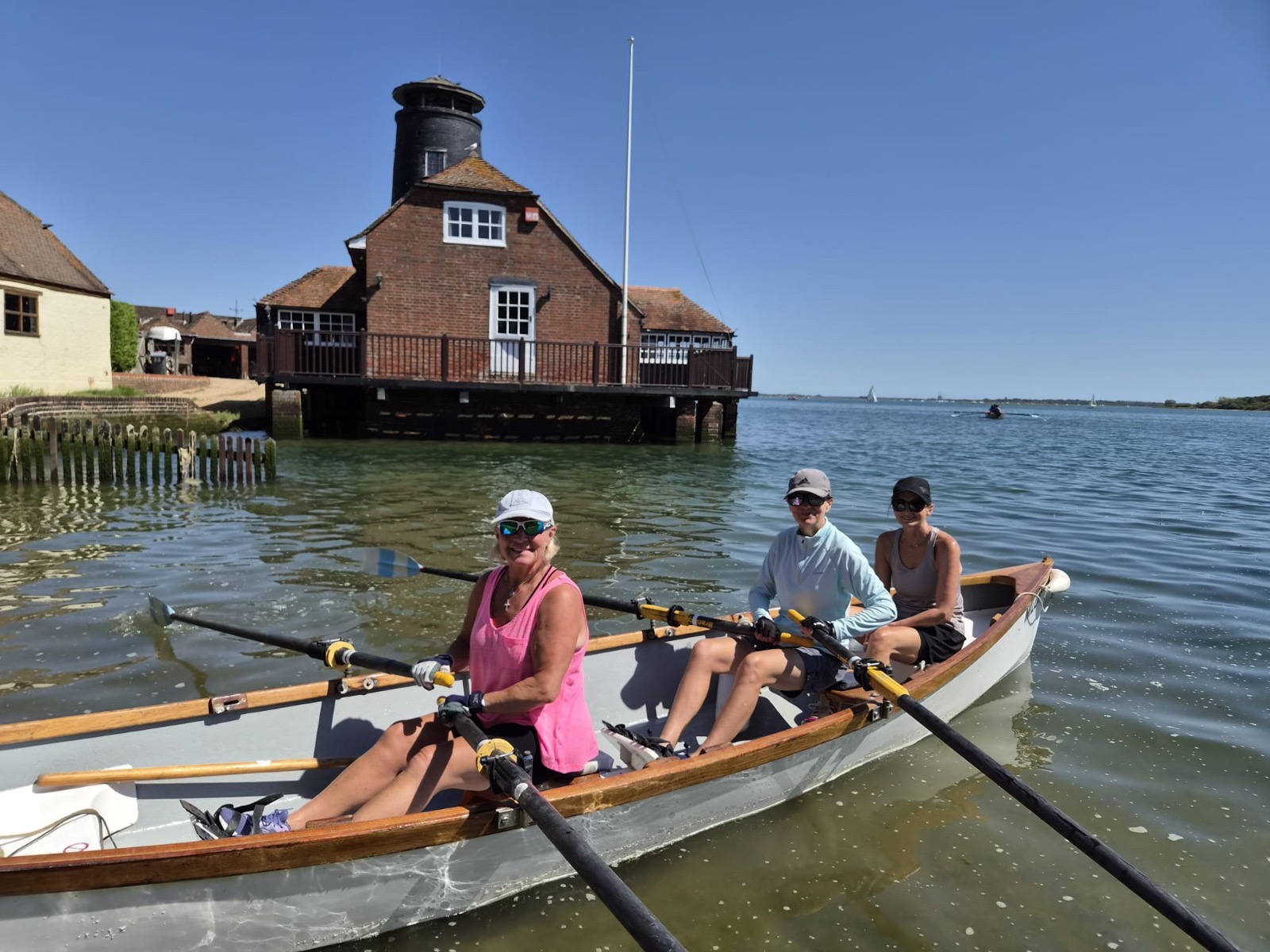 Three people rowing past a brick building on piles, with open harbour water beyond.