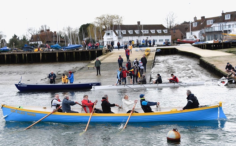 Rowers in a blue and yellow boat beside a harbour slipway, with other crews and people gathered on the pontoon.