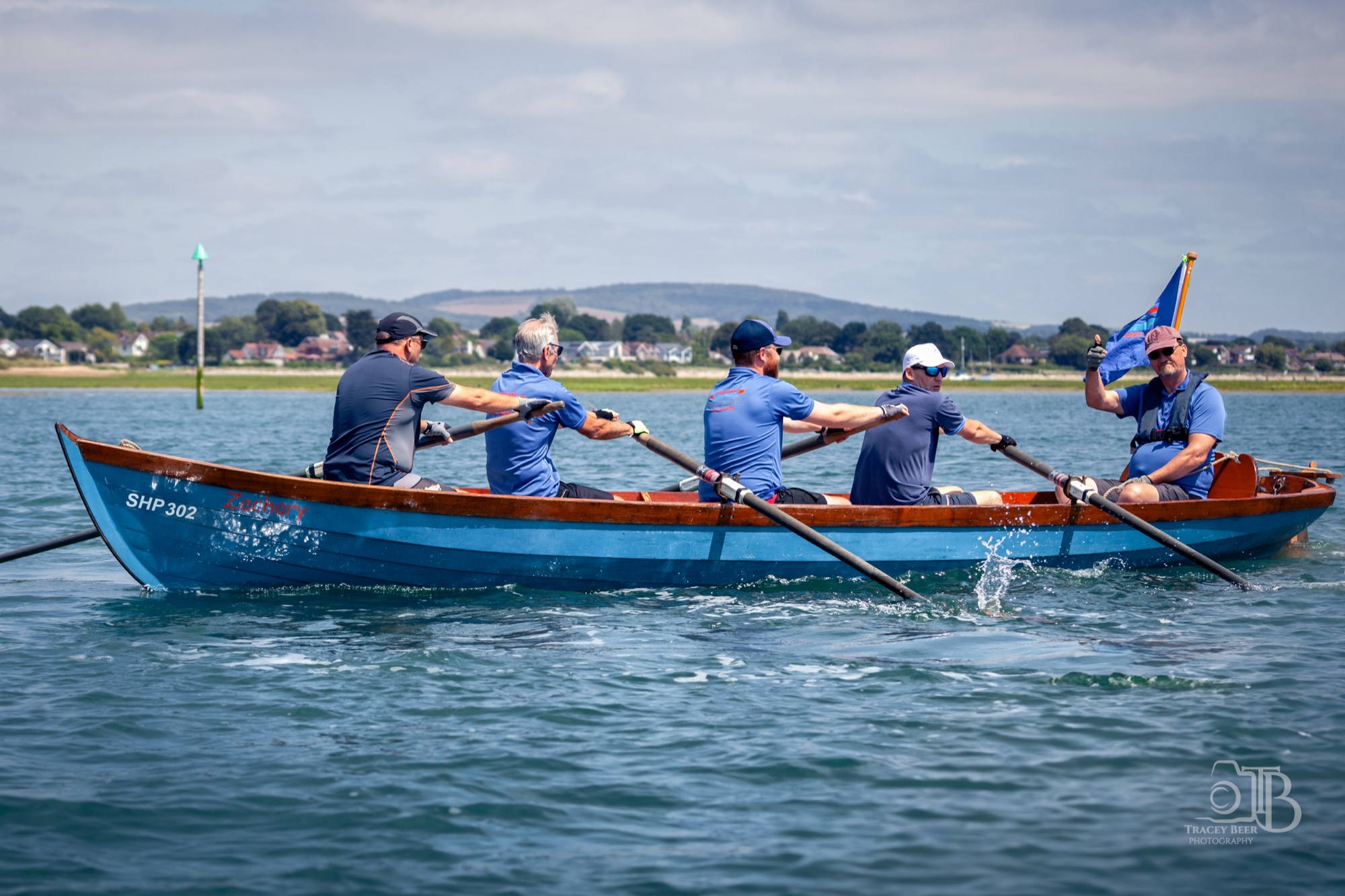 Five people rowing a blue fixed-seat boat on harbour water, one of them holding a blue flag.
