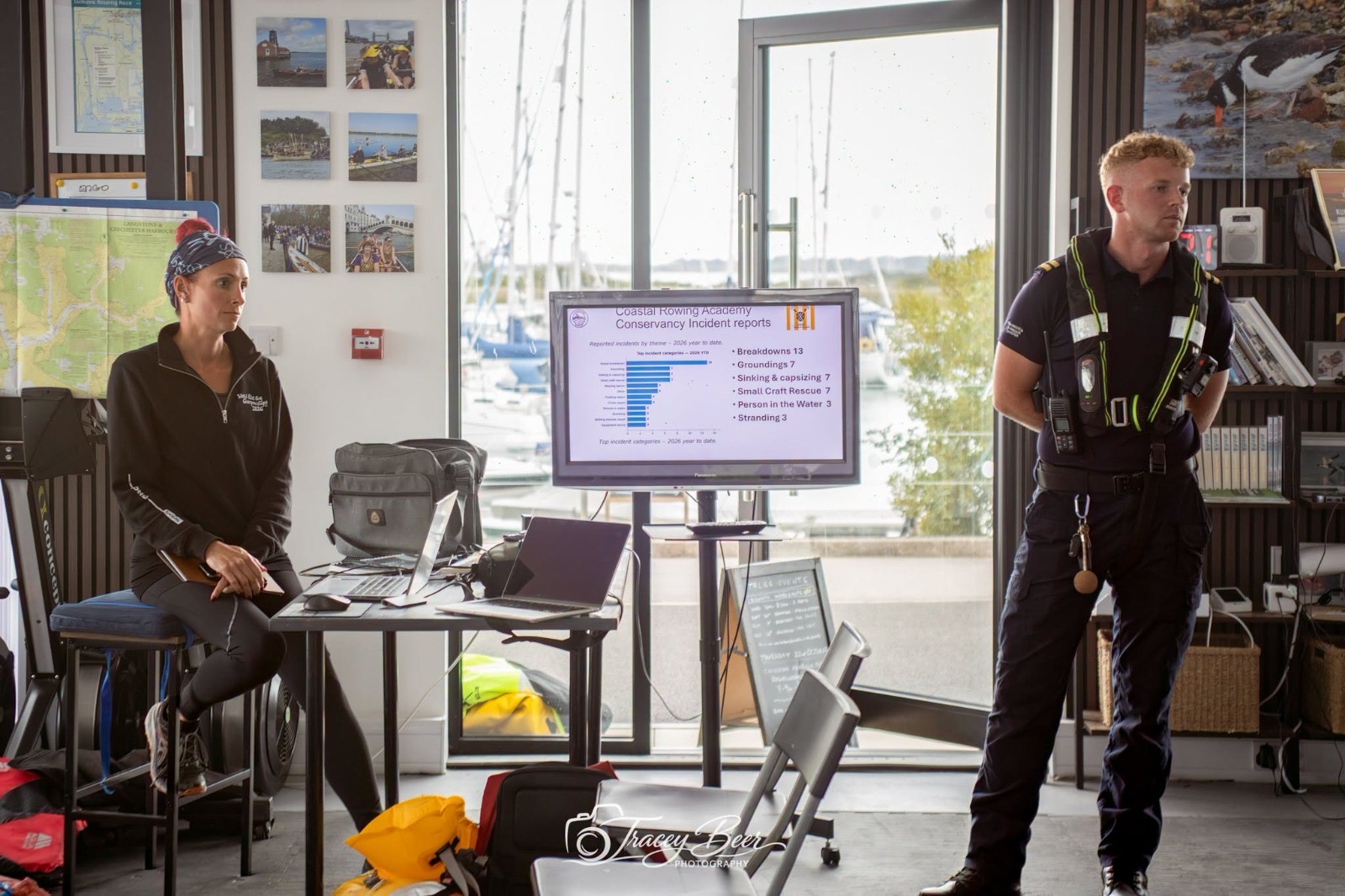 A harbour officer standing beside a seated woman during an indoor briefing, with the harbour visible through the windows.