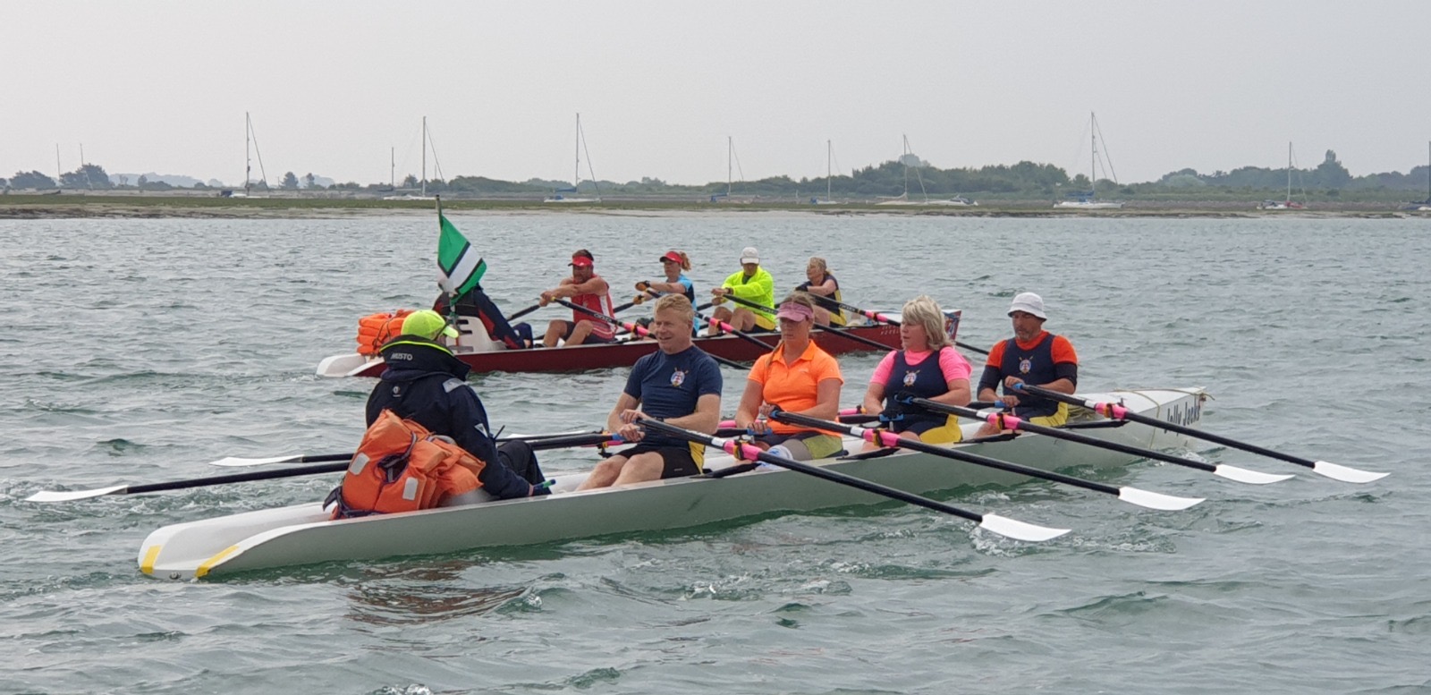 Rowers in a white coastal boat, with a red boat behind them and moored yachts along the far shore.